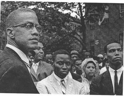 Malcolm X at an outdoor rally, a crowd of onlookers behind him, standing across from Louis Farrakhan at the far right. Photographer unknown.