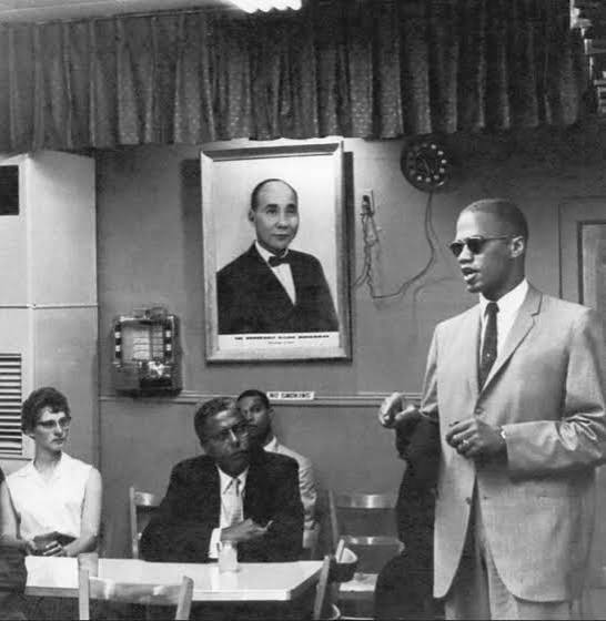 Malcolm X speaks beneath a portrait of "The Honorable Elijah Muhammad" at a Nation of Islam gathering, with Louis Farrakhan seated in the back row. Photographer unknown.