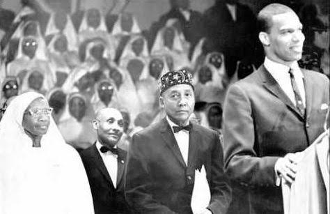 Elijah Muhammad, center, with Clara Muhammad, left, before Nation of Islam women in white at a gathering, with Louis Farrakhan at the podium, right. Photographer unknown.