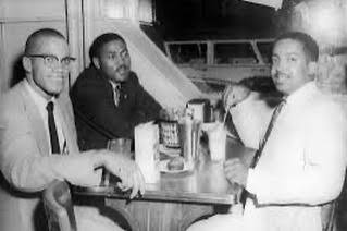 Malcolm X, left, seated at a restaurant table with Warith Deen Mohammed across from him and an unidentified man seated beside Malcolm. Photographer unknown.